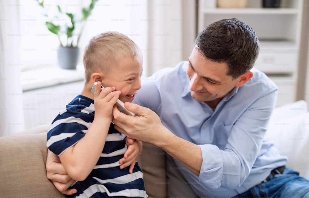 Un padre con un hijo feliz con síndrome de Down en el interior de su casa, usando un teléfono inteligente.