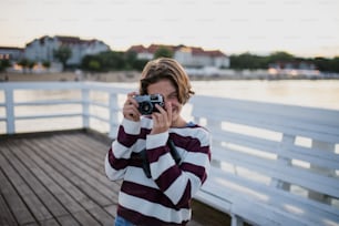 Preteen girl tourist taking a photo with camera on pier by sea at sunset, summer holiday concept.