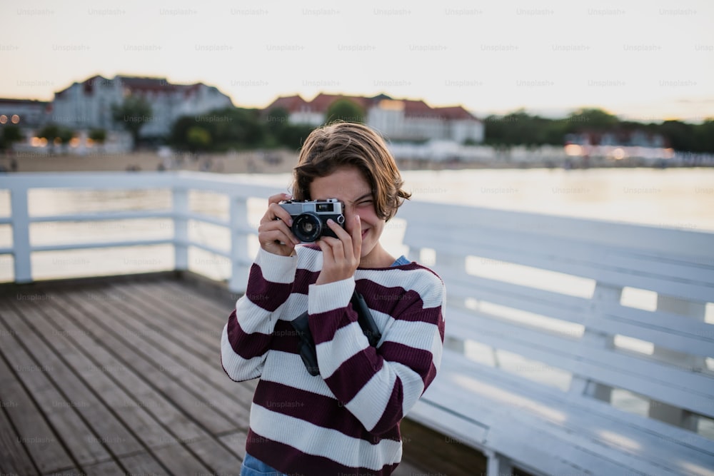 Turista della ragazza preadolescente che scatta una foto con la macchina fotografica sul molo dal mare al tramonto, concetto di vacanza estiva.