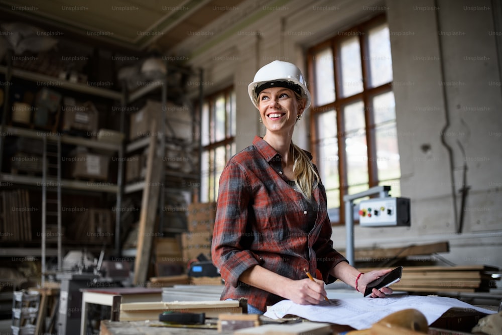 Una ingeniera sosteniendo planos y un teléfono inteligente en el interior de un taller de carpintería.