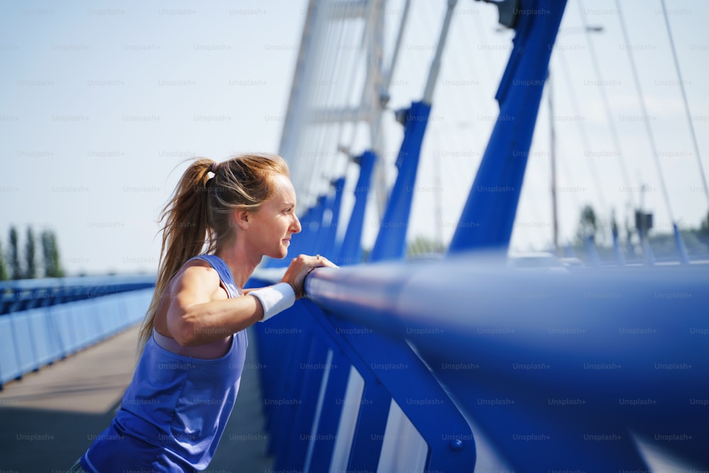 Uma corredora adulta média que se estende ao ar livre na ponte na cidade, conceito de estilo de vida saudável.