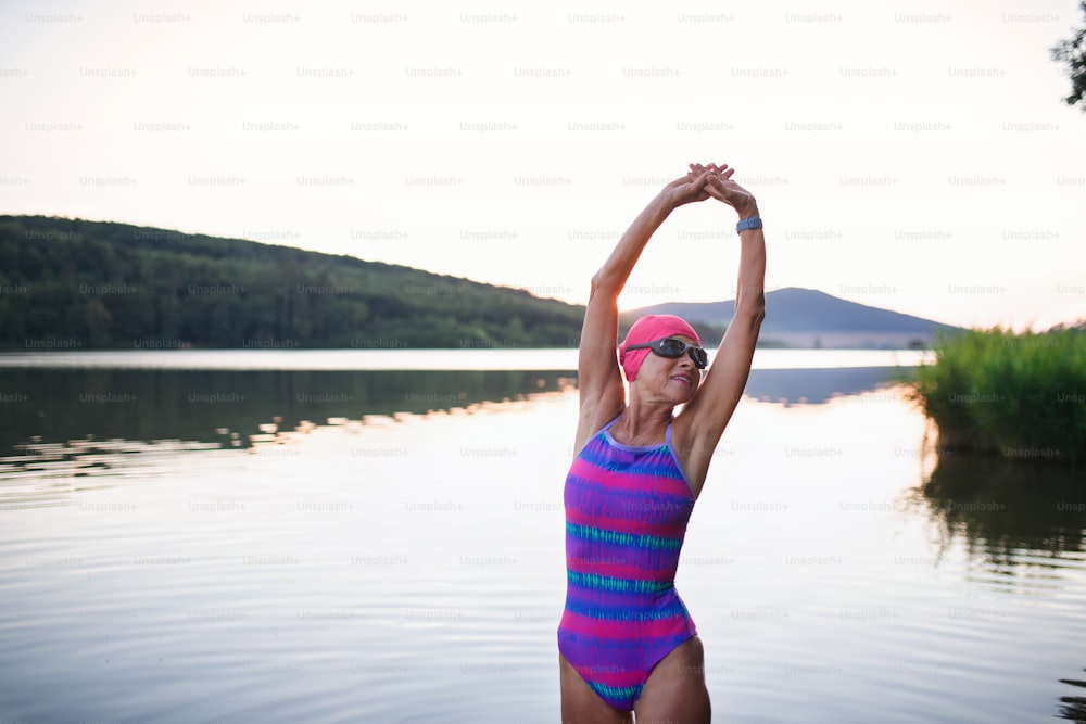 Un portrait d’une nageuse senior active debout et s’étirant à l’extérieur au bord d’un lac.