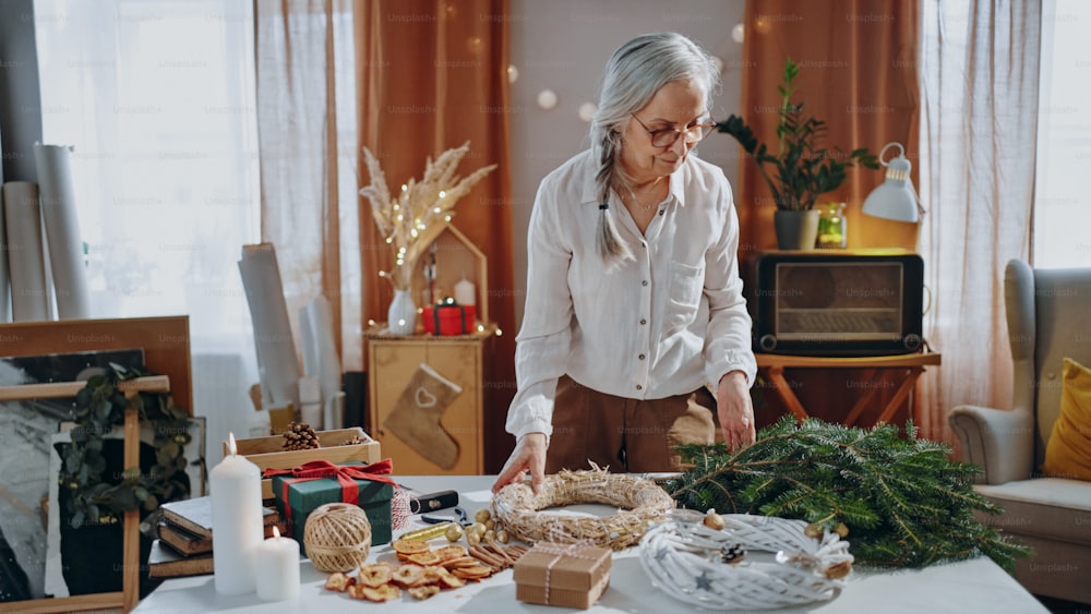 Mujer mayor que hace una corona de Navidad de materiales naturales en el interior de casa