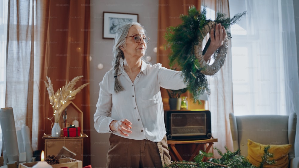 Mujer mayor que hace una corona de Navidad de materiales naturales en el interior de casa