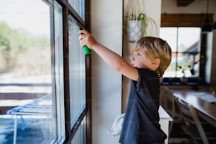 Vista lateral de um menino limpando janelas dentro de casa, conceito de tarefas diárias.