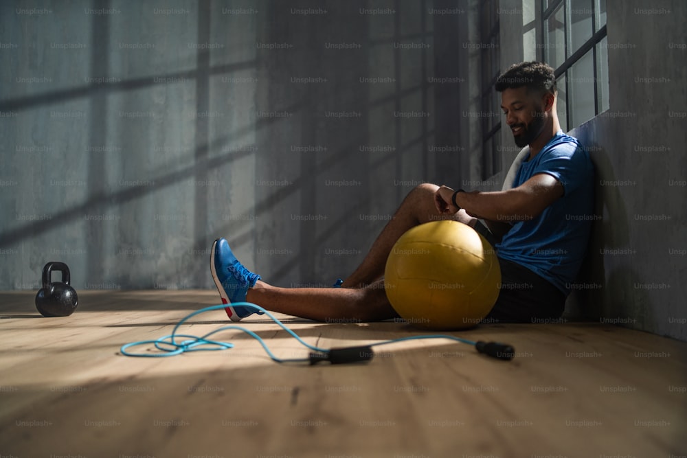 A young African American sportsman sitting and using smartwatch indoors in gym, workout training concept.