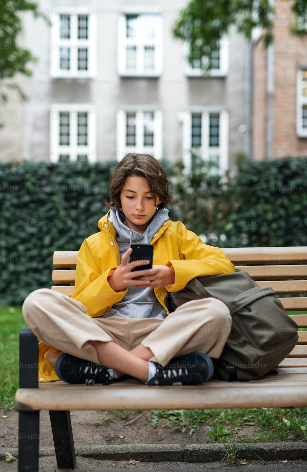 A preteen schoolgirl sitting on bench and using smartphone outdoors in town.