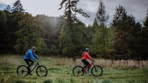 A side view of active senior couple riding bikes outdoors in forest in autumn day.