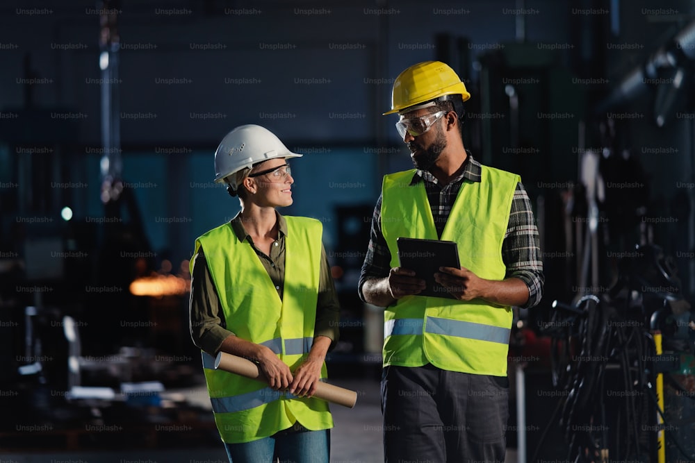 A portrait of industrial inspectors with tablet doing a general check up and talking indoors at metal workshop.