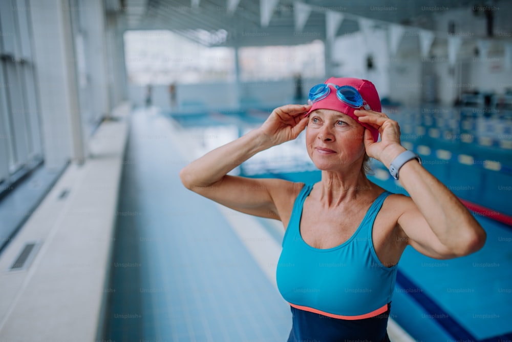 A senior woman looking at watch and putting on goggles before swim in indoors swimming pool.