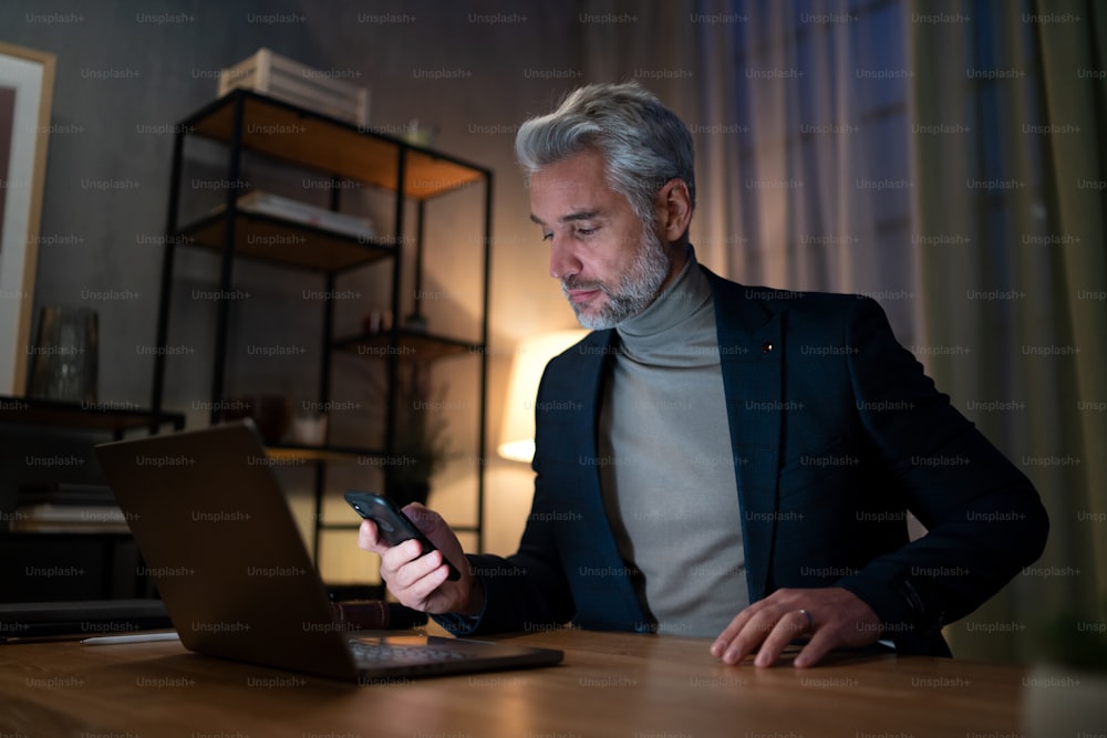 A mature businessman working on laptop at desk indoors in office at night.