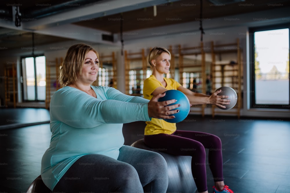 Una mujer feliz con sobrepeso sentada en la pelota fintess y haciendo ejercicio con el entrenador personal en el gimnasio