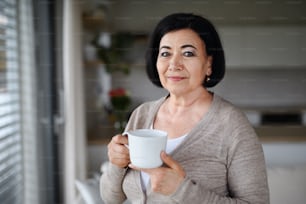 A happy senior woman with coffee standing and looking at camera indoors at home.