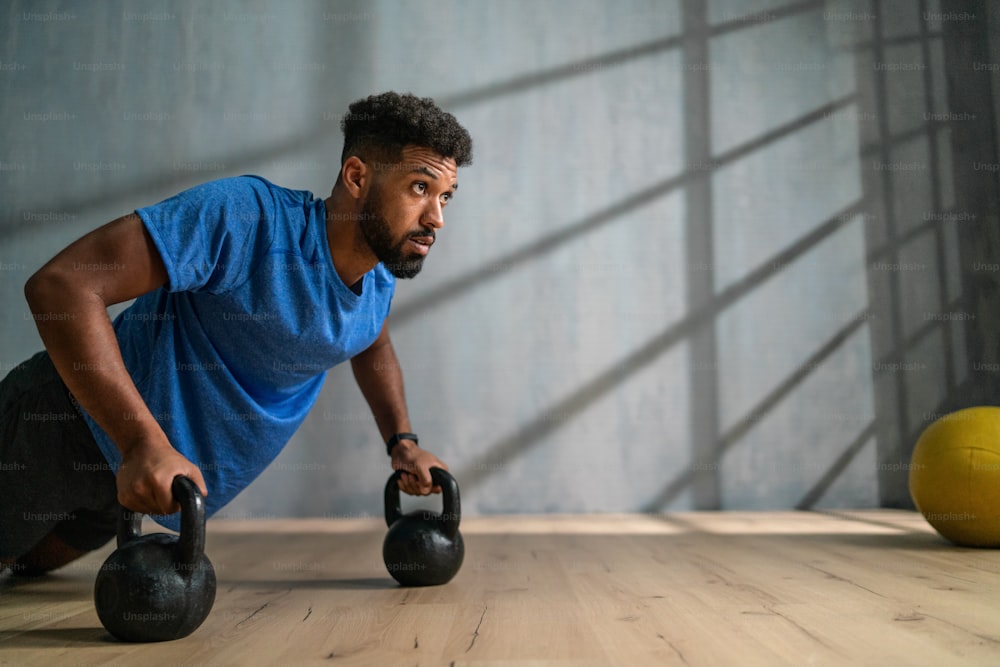 A young African American sportsman doing push ups with kettle bells indoors, workout training concept.