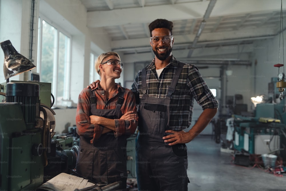 A portrait of young biracial industrial colleagues working indoors in metal workshop, smiling and looking at camera.