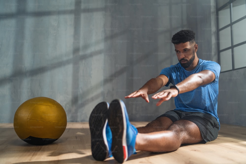 Un joven deportista afroamericano sentado en el suelo y haciendo ejercicios de estiramiento en interiores, concepto de entrenamiento de entrenamiento.