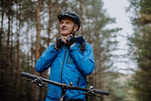 Un ciclista mayor activo de pie con una bicicleta al aire libre en el bosque en un día de otoño.