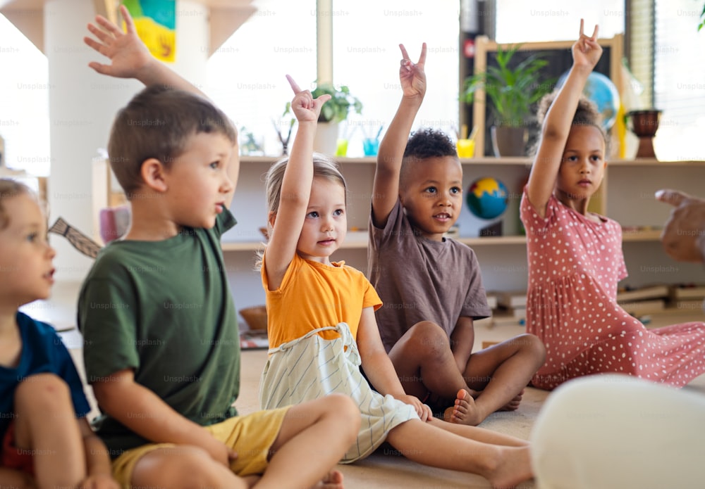 Um grupo de pequenas crianças da creche sentadas no chão dentro de casa de aula, levantando as mãos.