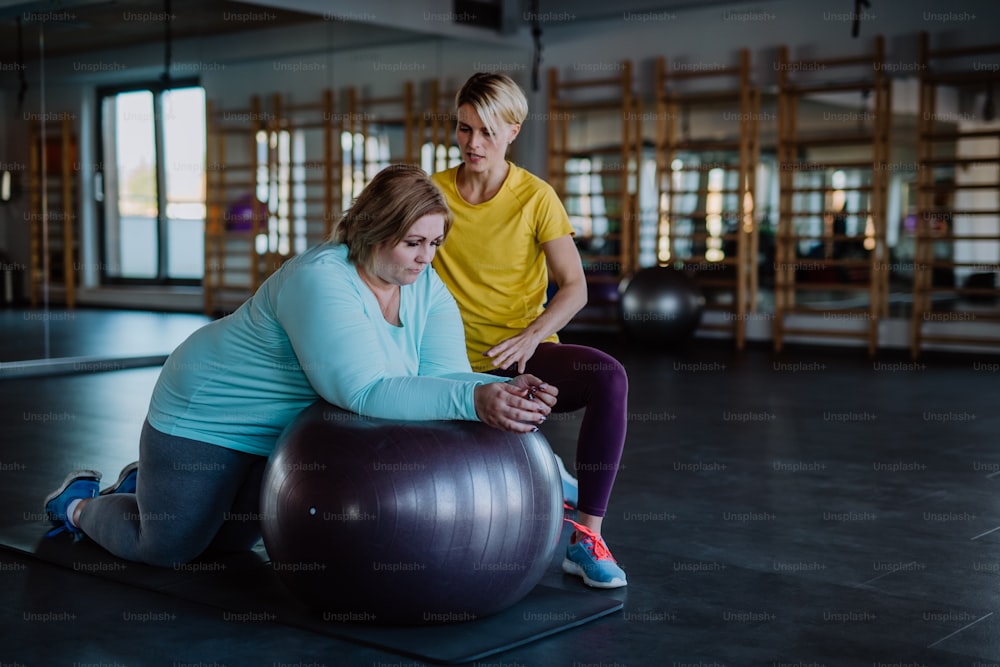A happy overweight woman exercising with personal trainer on fintess ball in gym