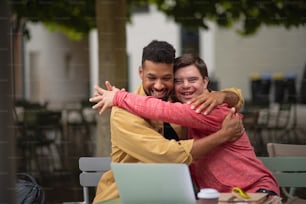 Happy young man with Down syndrome and mentoring friend with arms around outdoors in cafe using laptop