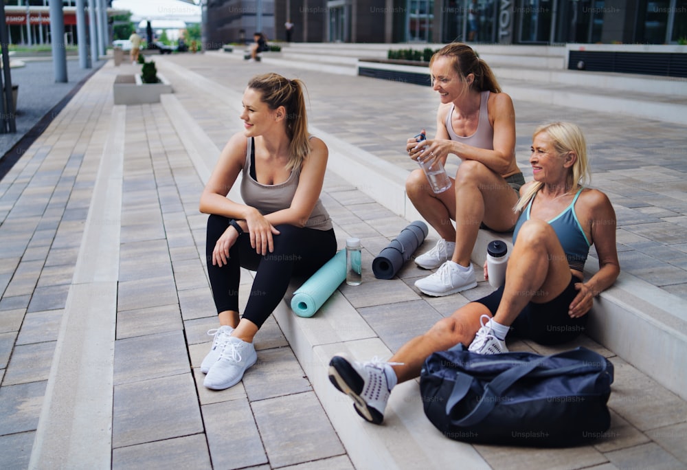 Um grupo de mulheres jovens e idosas sentadas após o exercício ao ar livre na cidade, conversando.