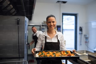 A happy female cook looking at camera and holding tray with baked pumpkin pieces in commercial kitchen.