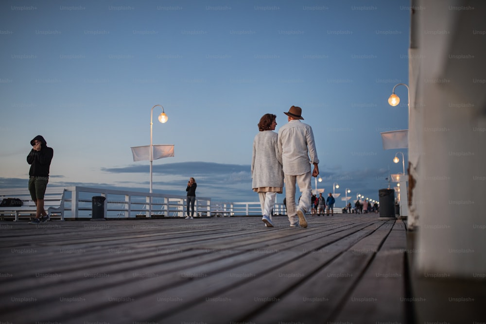 Eine rückwärtige niedrige Winkelansicht eines glücklichen älteren Paares, das in der Abenddämmerung auf dem Pier am Meer im Freien spazieren geht und Händchen hält.