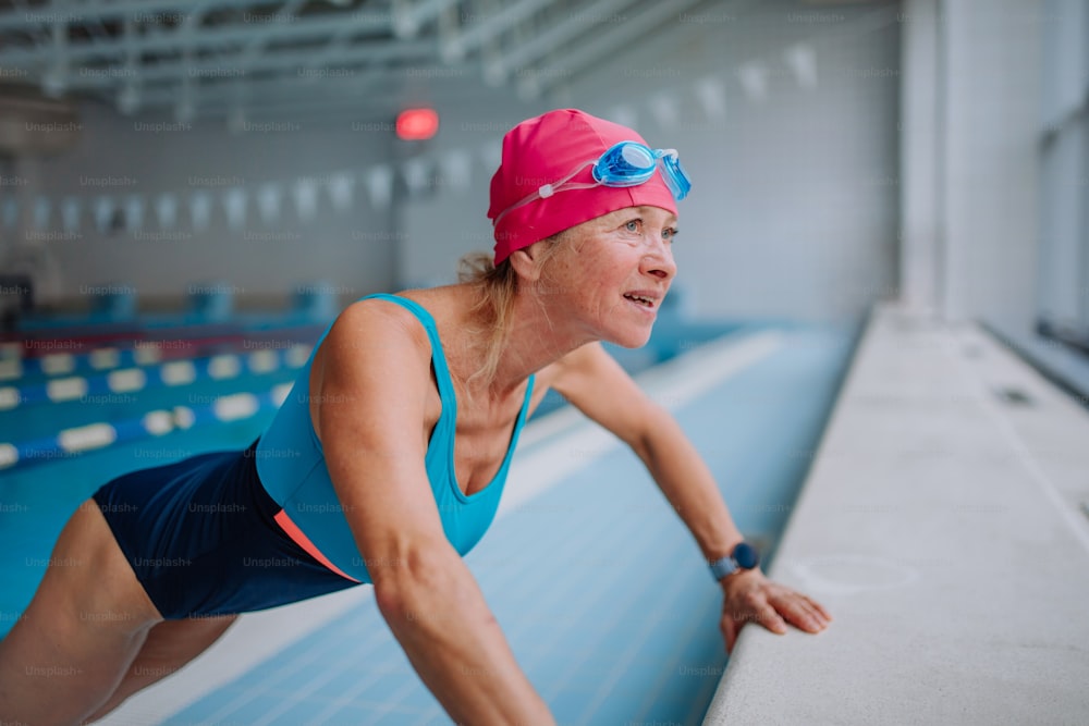 An active senior woman doing warming up exercise indoors in public swimming pool.