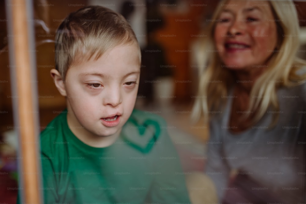 A boy with Down syndrome with his grandmother looking through window at home.