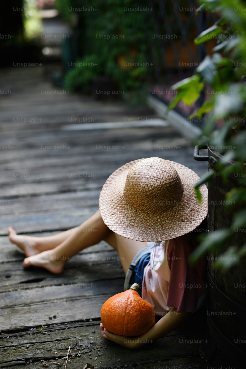 Hochwinkelansicht eines nicht erkennbaren kleinen Mädchens mit Hut, das draußen auf dem Bauernhof sitzt und Kürbis hält.