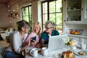 Amigos mayores felices usando una computadora portátil y un teléfono inteligente en el interior de casa, a la hora del café.