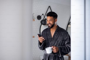 自宅のバスルームでコーヒーとバスローブを室内に着けている若い男性、朝のルーティンのコンセプト。
