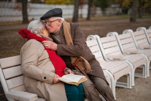 A happy senior couple sitting on bench and hugging outdoors in town park in autumn.