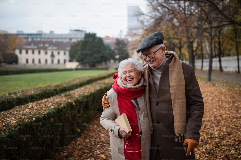 A happy senior couple on walk outdoors in town park in autumn, embracing and laughing.