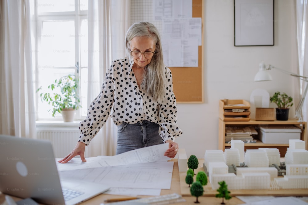 A senior woman architect with model of houses looking at blueprints in office.