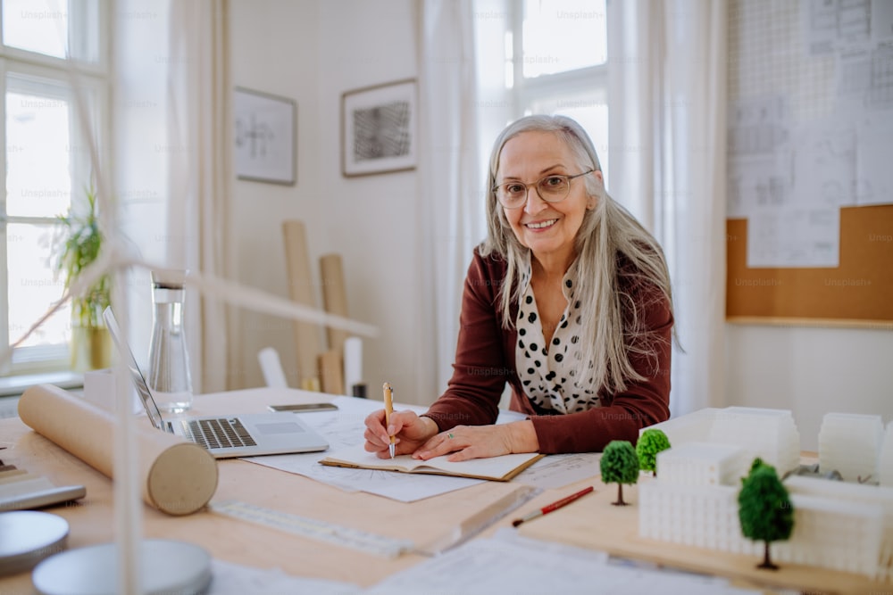 A senior woman architect with model of houses in office writing notes.