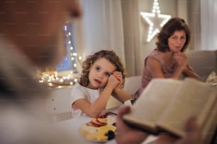 Small girl with parents and grandparents indoors celebrating Christmas, reading Bible.