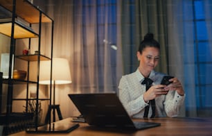 Eine glückliche mittelerwachsene Geschäftsfrau mit Tagebuch, die nachts im Büro am Laptop arbeitet und in die Kamera schaut.