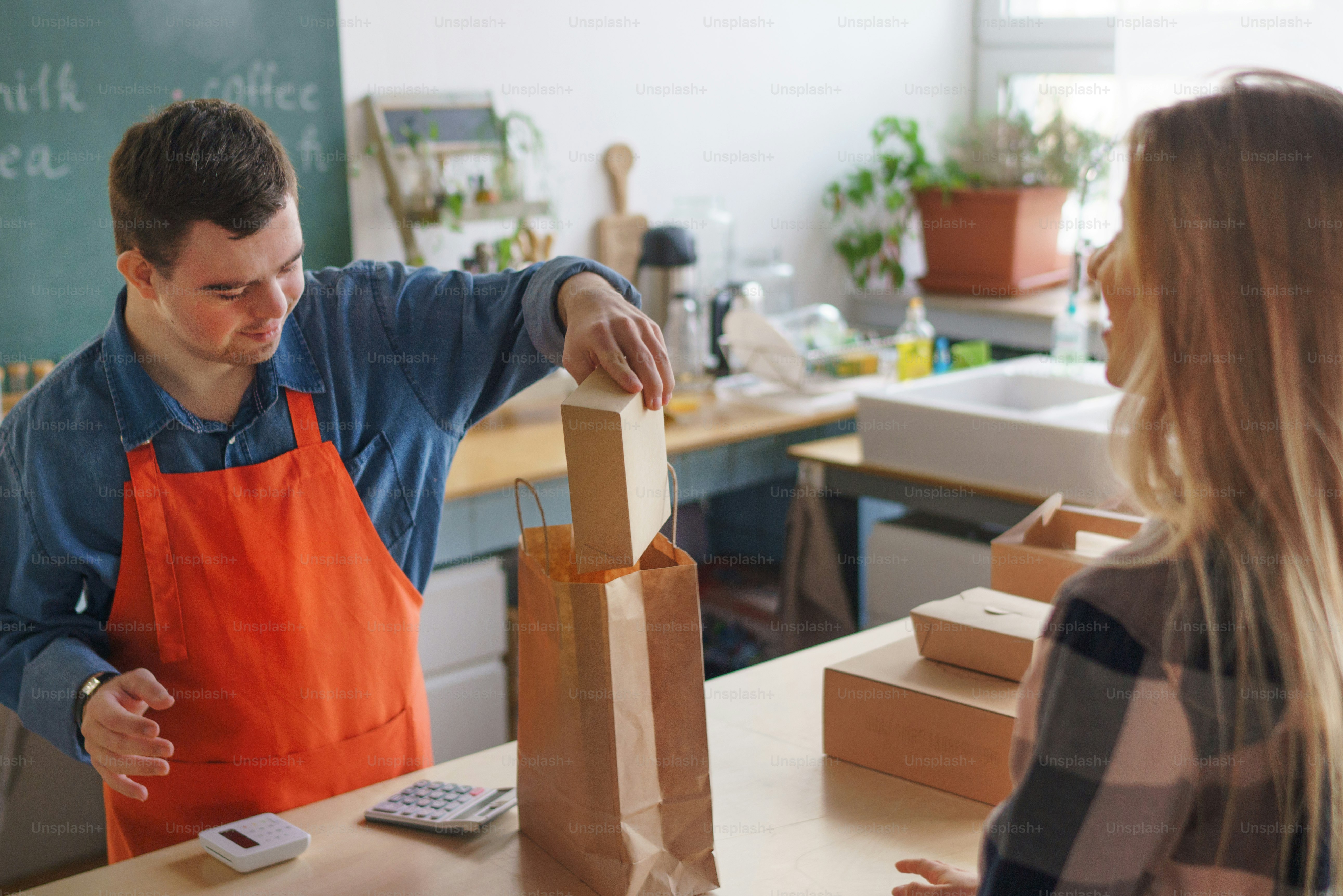 Un joven y alegre camarero con síndrome de Down que atiende a un cliente en un restaurante para llevar, concepto de inclusión social.