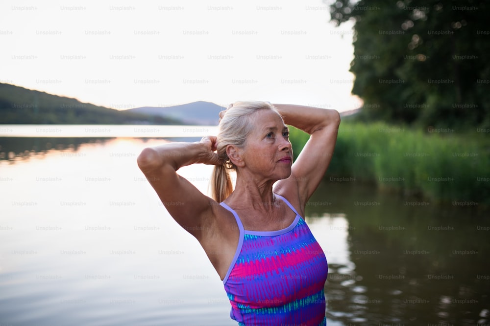 Un portrait d’une nageuse senior active debout et s’étirant à l’extérieur dans un lac.