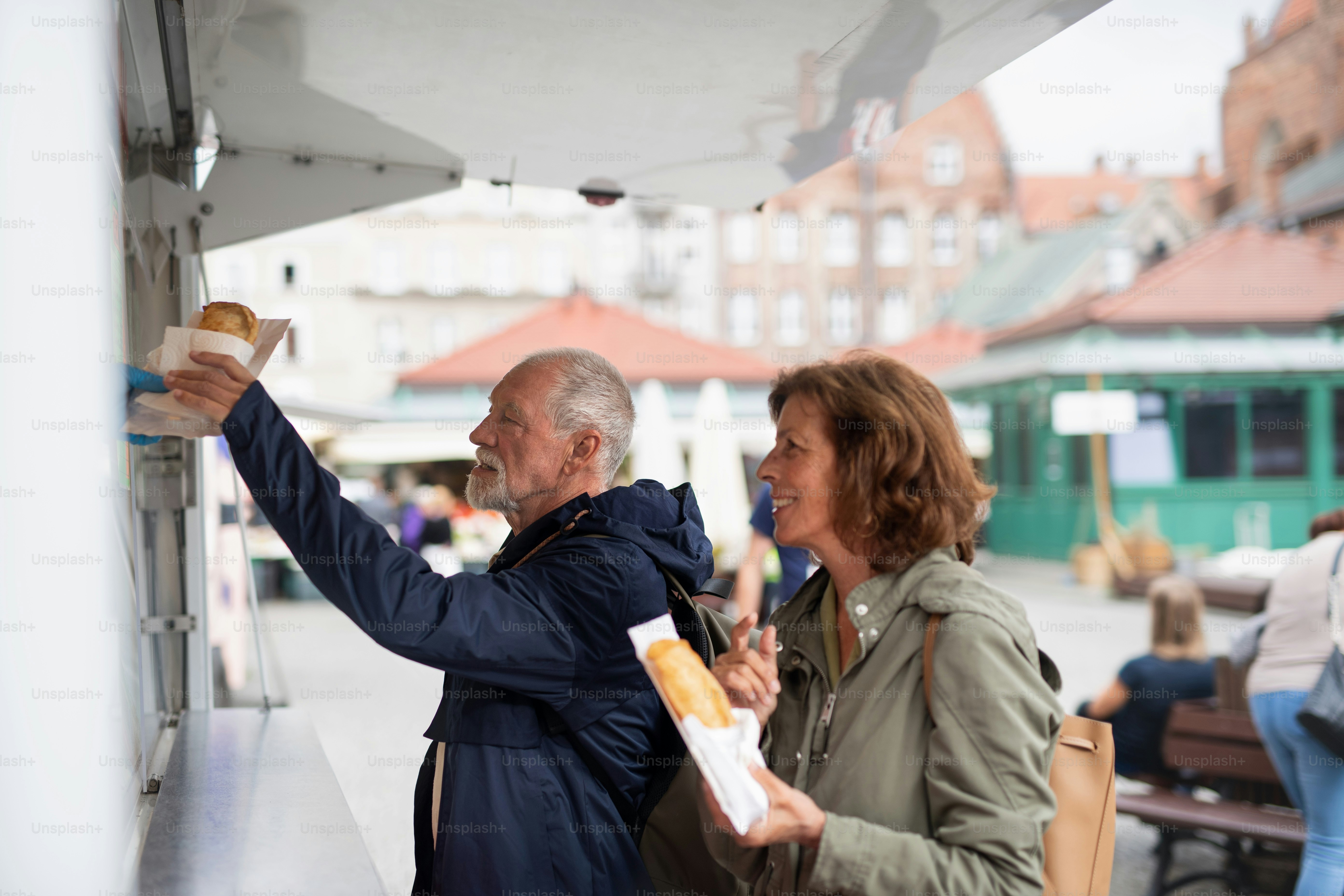 A happy senior couple tourists buying snack outdoors in street.