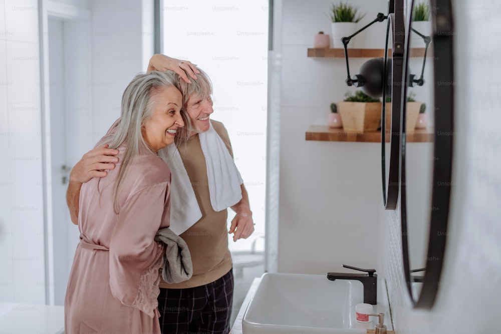 A senior couple in love in bathroom, looking at mirror and having fun, morning routine concept.