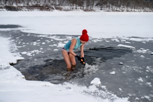 Mulher sênior ativa feliz em maiô quebrando gelo com machado ao ar livre no inverno, conceito de terapia fria.