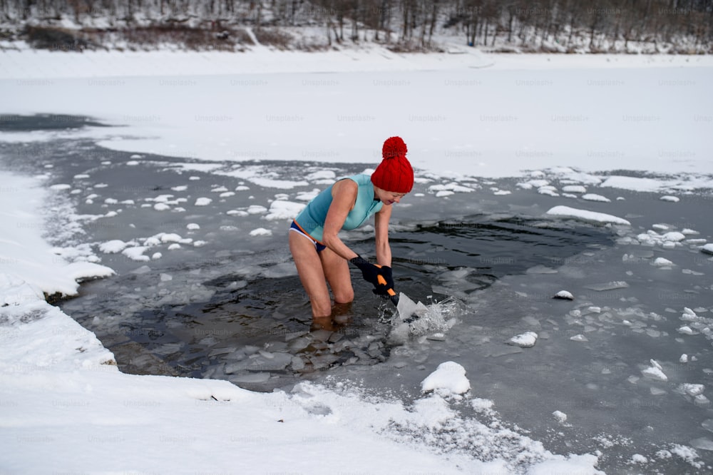 Glückliche aktive Seniorin im Badeanzug, die im Winter Eis mit Axt im Freien bricht, Kältetherapiekonzept.