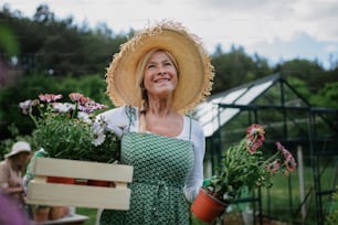Una florista mayor que lleva una caja con flores plantadas al aire libre en el jardín.