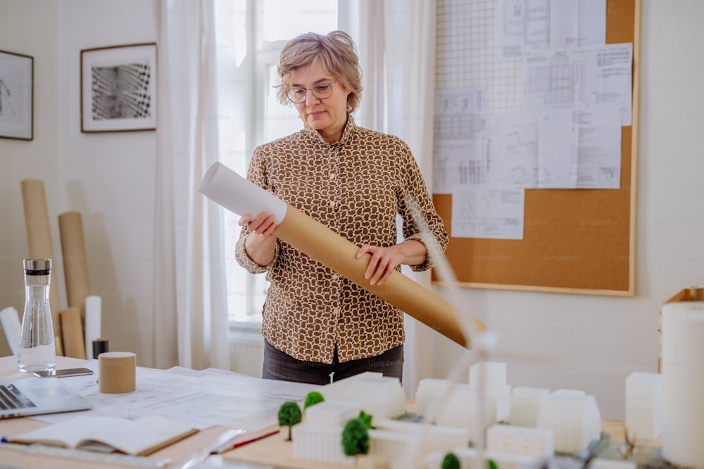 A mature woman architect pulling rolled-up blueprints out of tube in office.