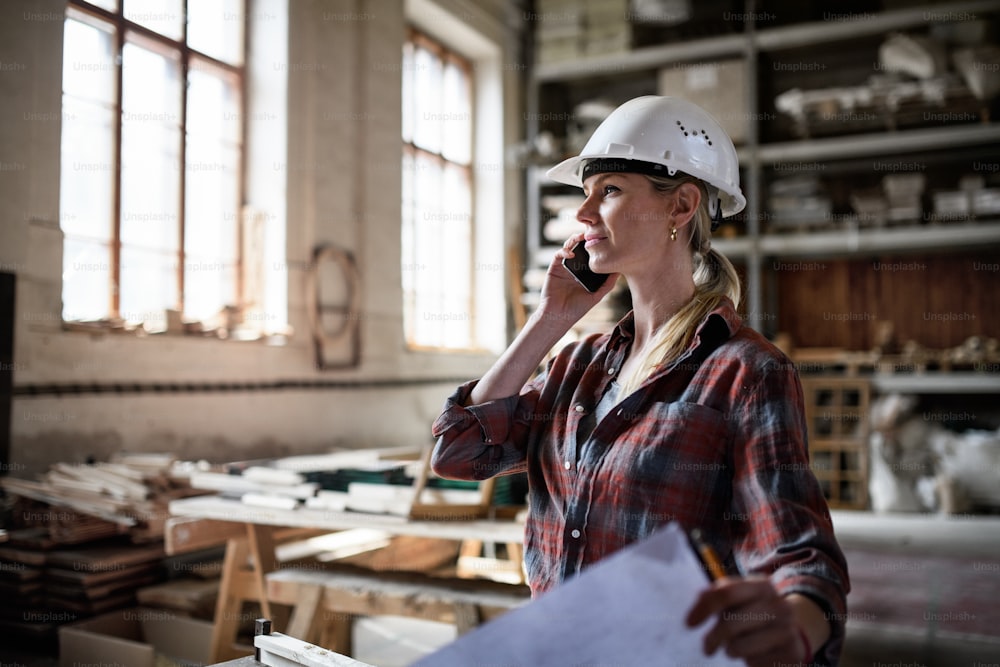 A female engineer holding blueprints and making phone call indoors in carpentry workshop.