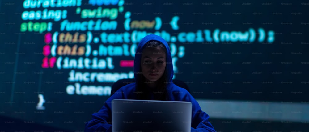 A hooded anonymous hacker woman by computer in the dark room at night, cyberwar concept.