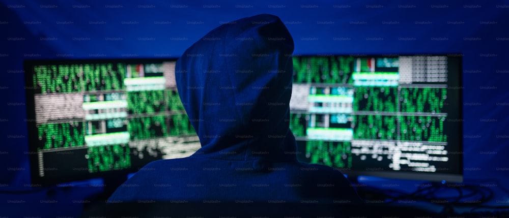 Una vista posteriore di un hacker incappucciato dal computer nella stanza buia di notte, concetto di guerra cibernetica.