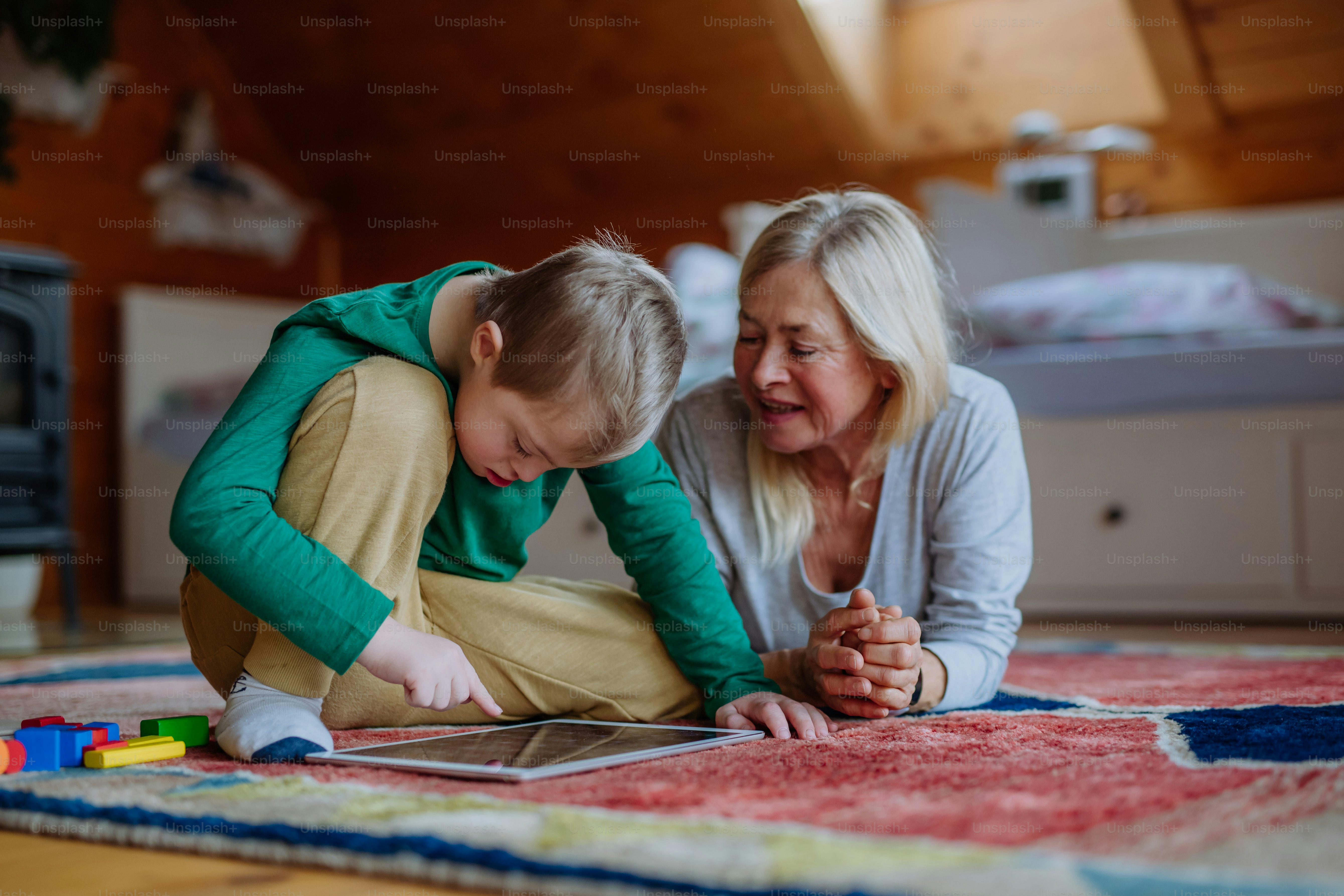 A boy with Down syndrome with his grandmother sitting on floor and using tablet at home.